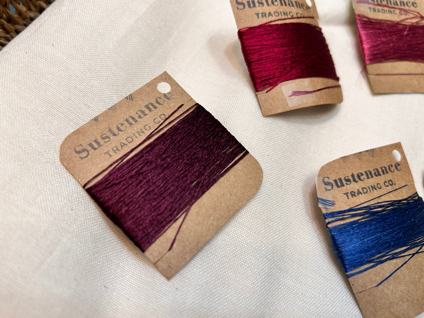 Bundles of red and blue thread on Sustenance Trading Co. cards on a white fabric background