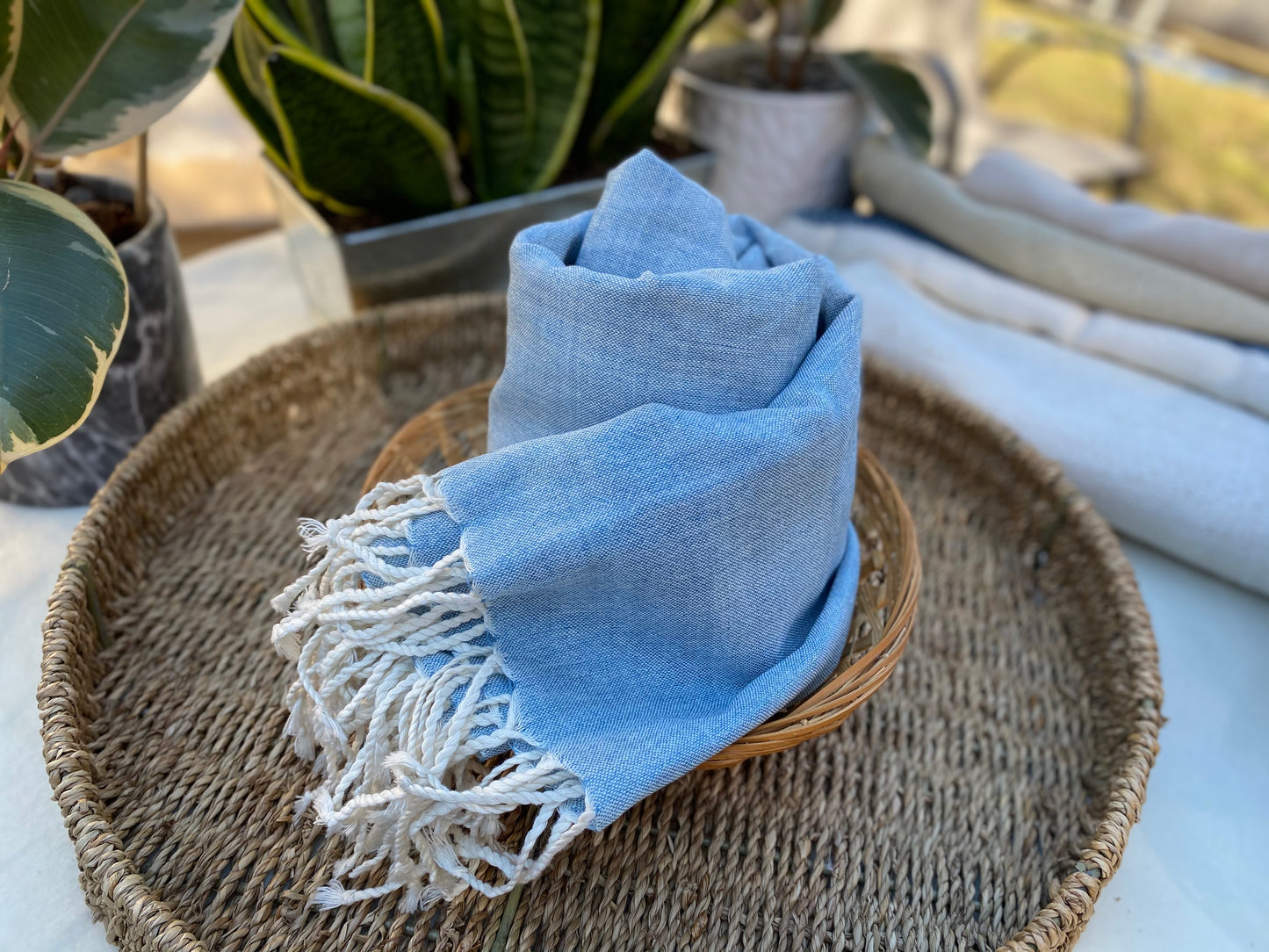 A light blue coloured scarf with off-white tassels is wound up in a roll on a flat basket.